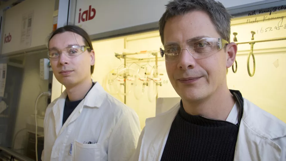 Två manliga forskare i vita rockar framför ett dragskåp.