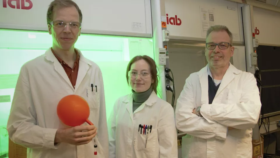 Tre kemiforskare i vita rockar framför ett dragskåp.