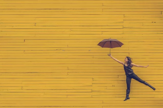 Kvinna med paraply hoppar med gul vägg i bakgrunden. Foto.