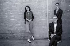 En kvinna och två män i kostym framför grå vägg.
