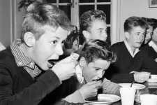 Barn som äter mat i en skolmatsal, med tallrikar och muggar framför sig. Svartvitt foto från slutet av 50-talet.