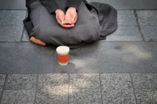 Person som sitter med benen i kors och knäppta händer vid en pappmugg på asfalterad gata. Foto.