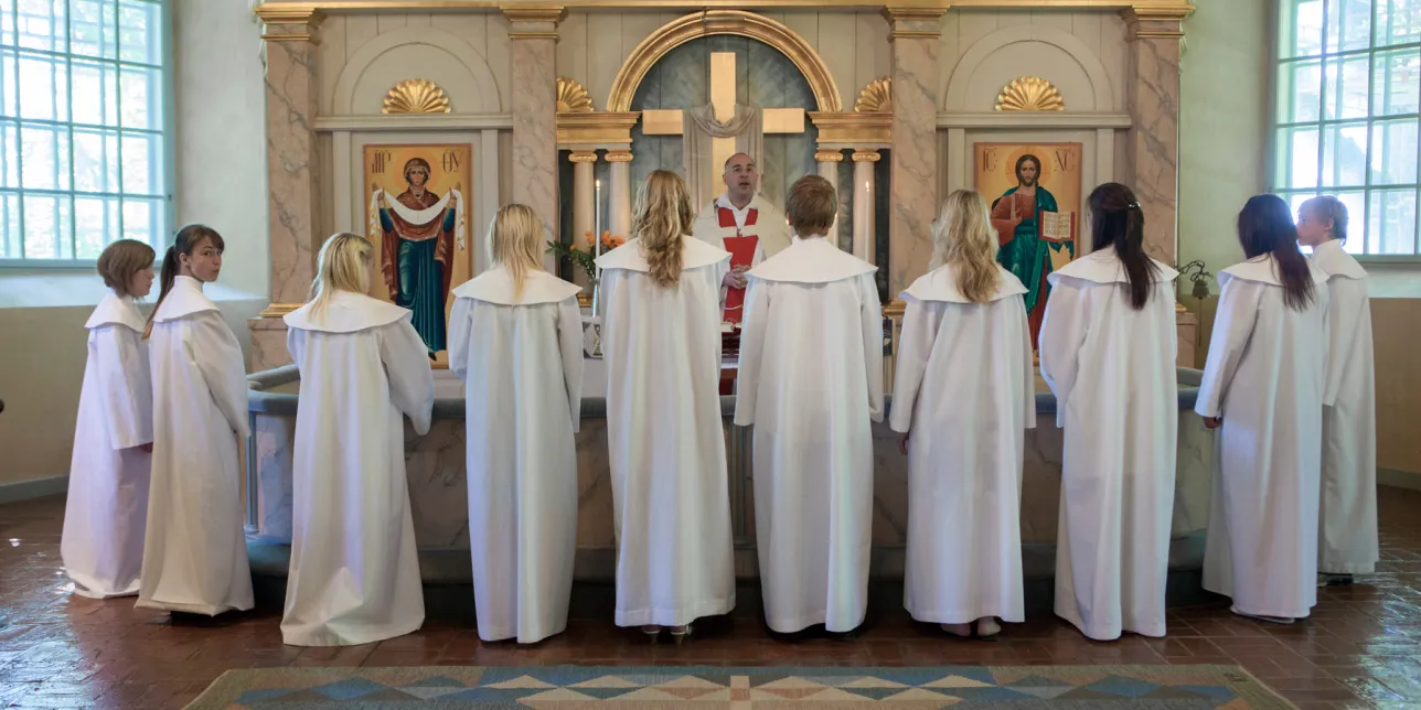Konfirmationsbild från Svenska kyrkan. Konfirmanderna står med ryggen mot försmlingen runt koret vända mot prästen som står med ryggen mot altaret.