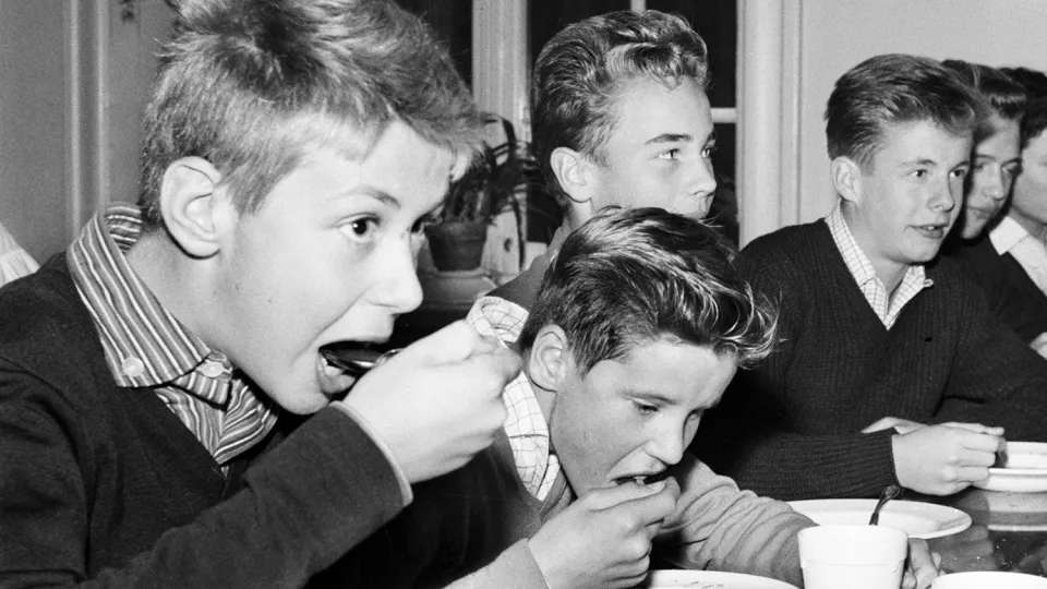Barn som äter mat i en skolmatsal, med tallrikar och muggar framför sig. Svartvitt foto från slutet av 50-talet.