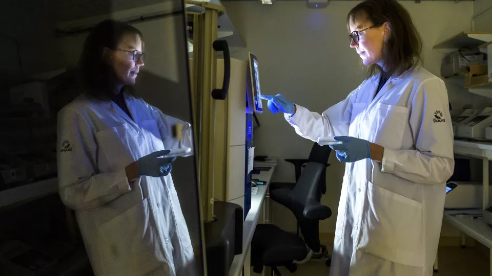 Forskare i vit labbrock tittar in i en maskin. Hennes spegelbild reflekteras i en glasruta. foto.