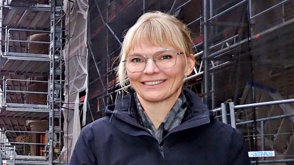 Kvinna i mörkblå jacka framför hus med byggnadsställningar. Foto.