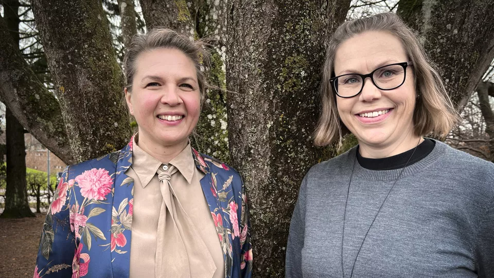 Foto. Kia Olsson och Ann-Catrin Johansson står bredvid varandra och lutar ryggarna mot ett träd. De ler och tittar in i kameran.