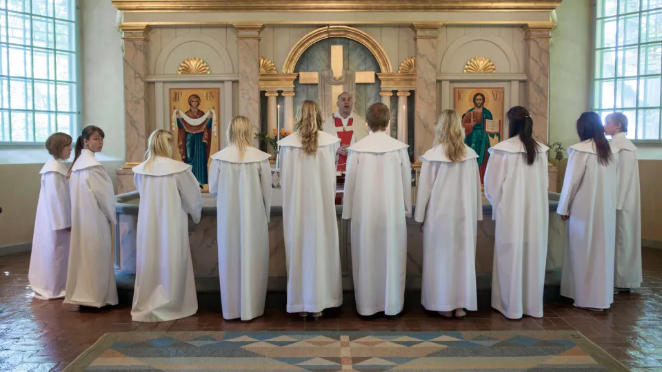 Konfirmationsbild från Svenska kyrkan. Konfirmanderna står med ryggen mot försmlingen runt koret vända mot prästen som står med ryggen mot altaret.