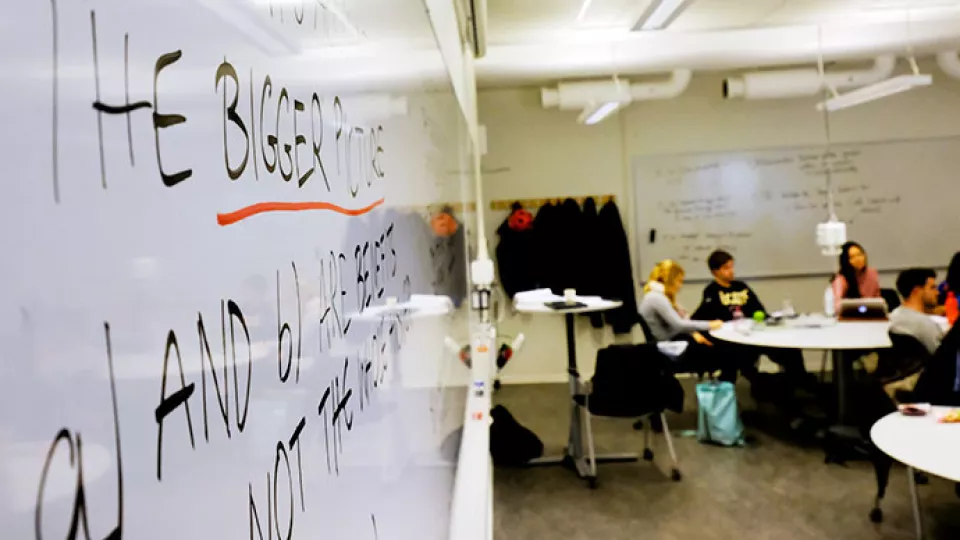 Detaljbild av en whiteboard, med studenter sittande runt bord i bakgrunden. På tavlan står det "the bigger picture". Foto.