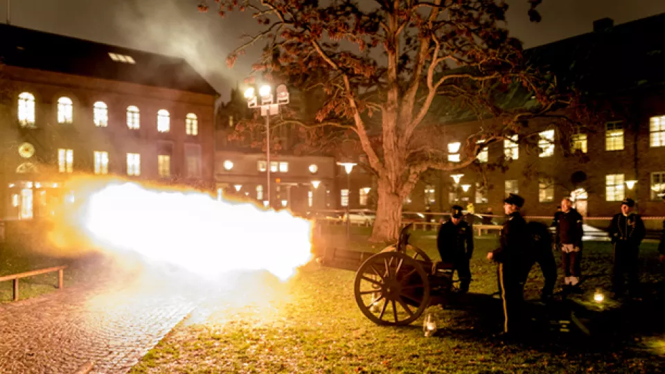 En kanon brinner av med eldsflamma i Lundagård. Fyra män i uniform står vid kanonen.