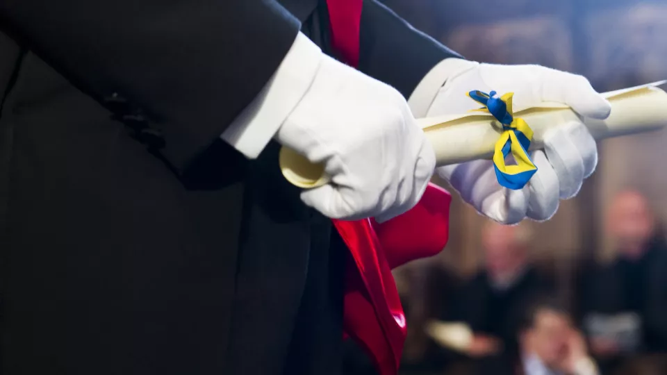 Närbild på händer med vita handskar som håller rullat diplom med blågult band