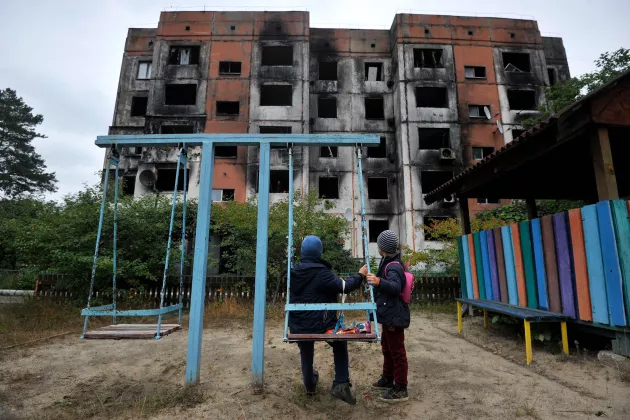  Barn står på en lekplats framför en förstörd byggnad i Kalynivka, norr om Kiev. Foto: Sergei Chuzavkov/AFP.
