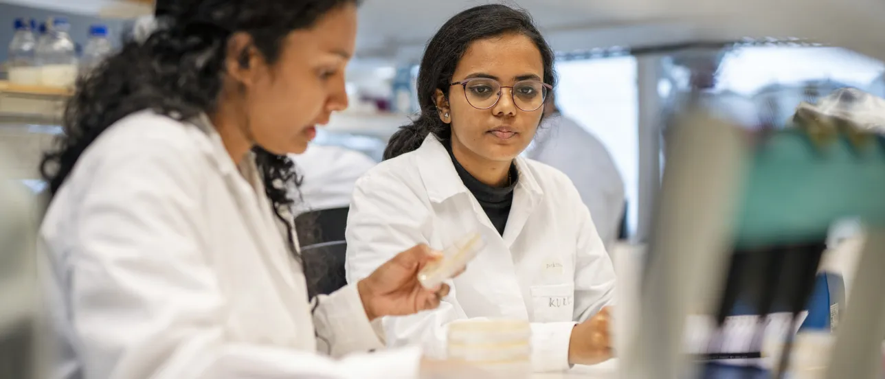 Två studenter i labbrockar diskuterar.