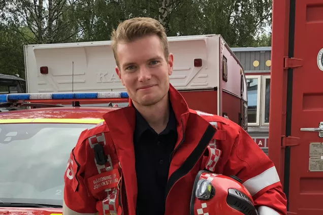 Johan står vid en brandbil i brandmansutrustning och ler. Foto.