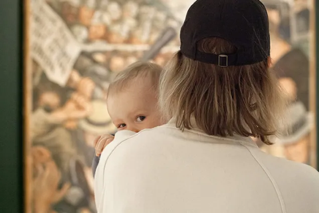 Ett litet barn kikar fram bakom en förälders axel och tittar in i kameran. Föräldern har ryggen mot kameran och tittar på en målning.