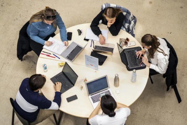 Fem studenter med laptops vid runt bord. Foto.