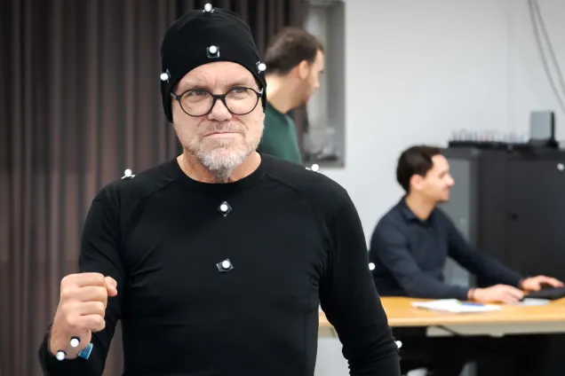 Person i svart dräkt med rörelsesensorer, med personer som arbetar vid ett skrivbord i bakgrunden. Foto.