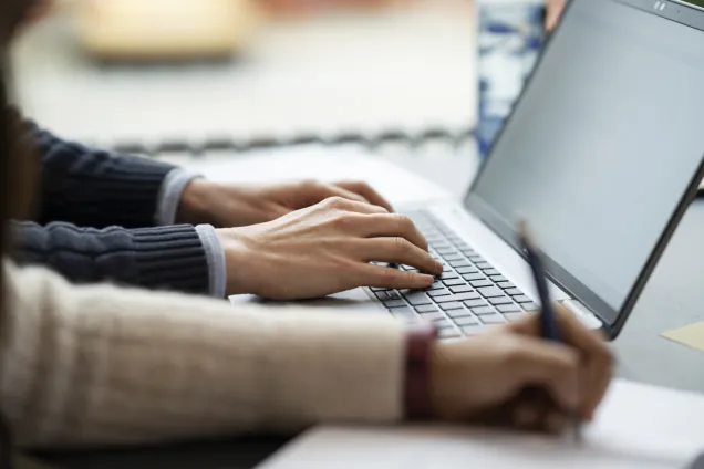 Två personer som sitter och skriver, en vars händer skriver på en laptop och en som skriver för hand. Foto.