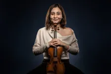 Ekon Kuzukiran håller violinen med båda händer. Hon har ljus skjorta och ett ler stort. 