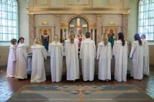 Konfirmationsbild från Svenska kyrkan. Konfirmanderna står med ryggen mot försmlingen runt koret vända mot prästen som står med ryggen mot altaret.