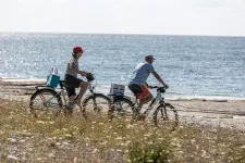 Ett seniorpar cyklar ned mot strandkanten. Blå himmel, strandlandskap. Foto: Carl Tronders/Unsplash 