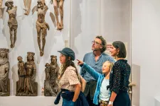En familj med två vuxna och två barn står och tittar på träskulpturer. Det yngsta barnet pekar upp på en skulptur. I bakgrunden kan man se att en liten tomtenisse gömmer sig bland skulpturerna.