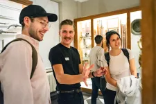 En leende manlig guide iförd svarta kläder håller en visning för en till vänster leende man iförd mörk keps, och en kvinna till höger iförd ryggsäck. I bakgrunden syns museimontrar.