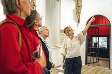 En pekande kvinna i vit blus och mörka byxor guidar och pekar med hela vänsterarmen uppåt, för att visa någonting för en grupp damer om tre som står till vänster om henne