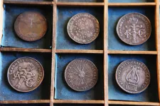 Sex stycken historiska mynt med bronsfärg ligger i två rader i små fack med turkos, sliten tygbakgrund