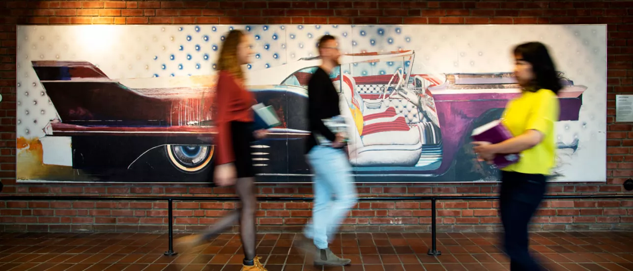 Tre personer i rörelse framför målning föreställande gammal amerikansk bil. Foto.
