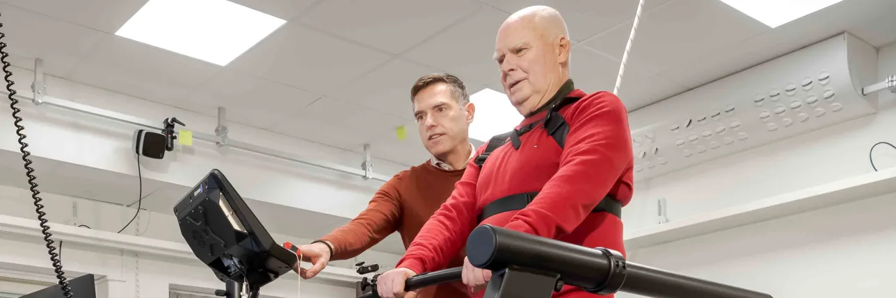 Person tränar på ett löpband med sele assisterad av en instruktör. Foto.