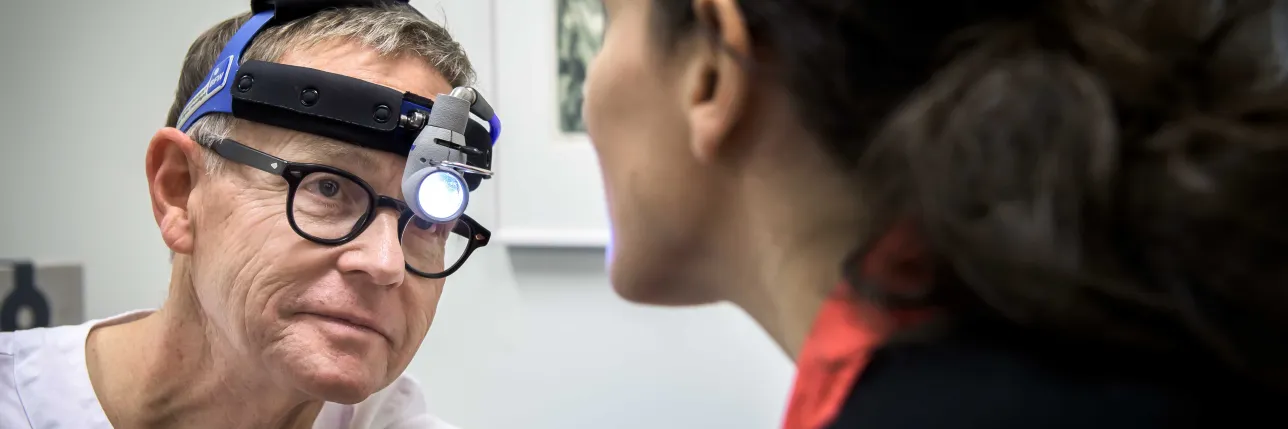 Person i vit vårduniform med ett ljusinstrument på huvudet. Foto.