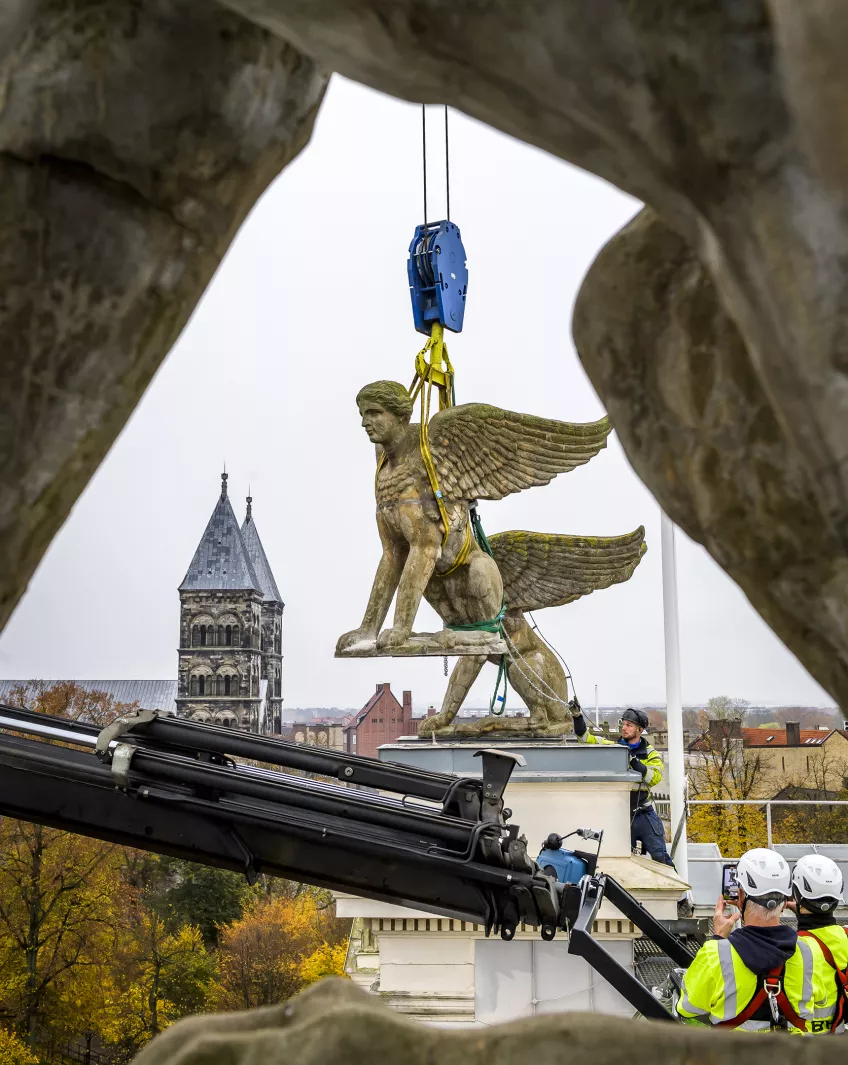 Sfinxerna lämnar nu universitetshusets tak, men kommer att ersättas under 2022. På bilden hänger sfinxen i luften med Lund som utsikt i bakgrunden.