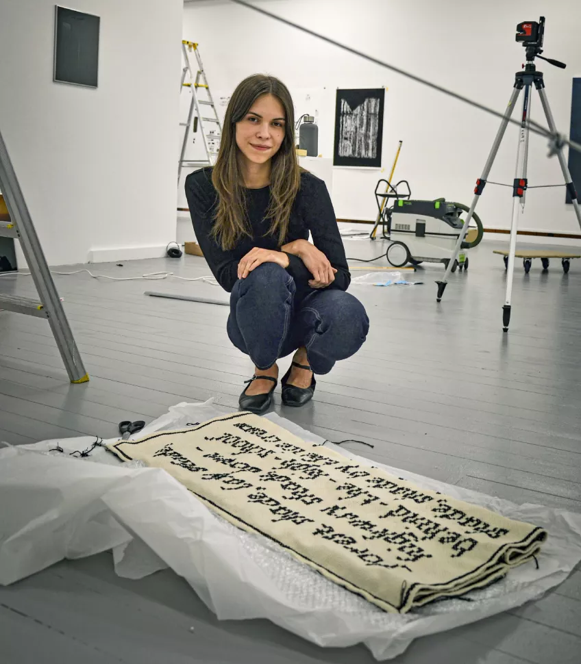 Foto. Konststudenten Liva Stare sitter på huk framför ett konstverk, ett broderat tygstycke.