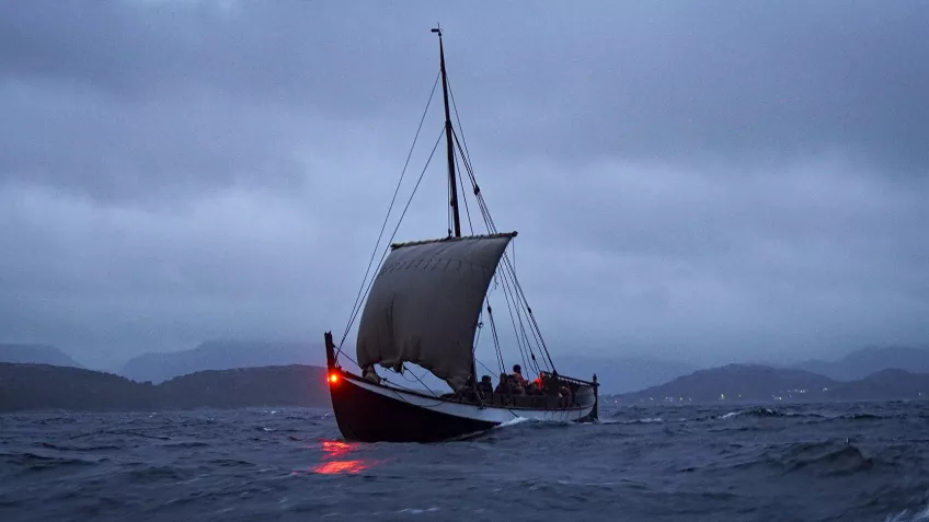 Foto av en båt, som liknar dåtidens vikingaskepp, ute till havs i kvällen. Seglet är hissat och i fören syns en röd lanterna.